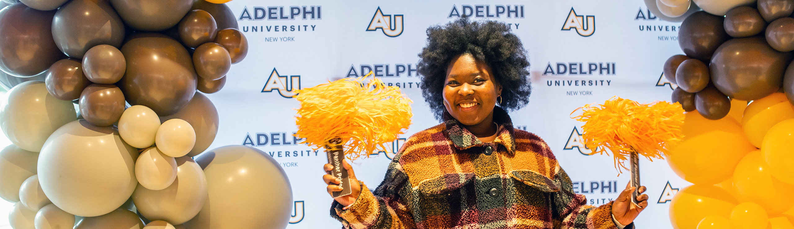 student smiling while holding poms in front of an Adelph iUniversity step-and-repeat